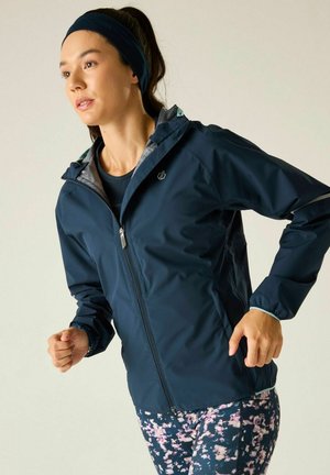 Vrouw die binnen jogt, gekleed in een marineblauwe jas met rits, een legging met bloemenpatroon en een donkere hoofdband.