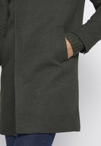 Manteau vert foncé en tissu texturé, avec une poche latérale et des poignets en tricot côtelé. Le design est simple et droit, renforçant sa polyvalence.