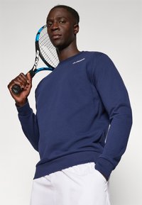 Marineblauwe sweatshirt van zachte stof, met een ronde halslijn en lange mouwen. Een tennisracket wordt in de linkerhand vastgehouden.