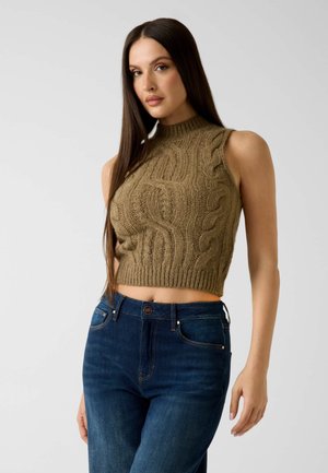 Donna con capelli lunghi e scuri che indossa un top corto marrone a maglia con motivo a trecce senza maniche e jeans a vita alta blu, su sfondo neutro.