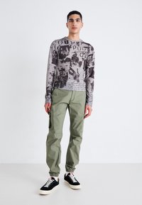 Camisa de manga larga con un patrón abstracto en negro y gris, combinada con pantalones cargo verde oliva con cremalleras laterales y zapatillas negras.