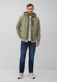 Veste zippée vert clair avec capuche, poches latérales et accents noirs, portée sur une chemise blanche, associée à un jean bleu et des baskets.