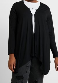 Osoba nosząca czarny kardigan z długim rękawem i otwartym przodem na białej koszulce z grafiką oraz zniszczonych czarnych dżinsach.