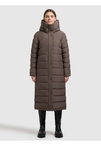 Geacă puf maro cu guler înalt, lungime până la genunchi, matlasată orizontal, închidere frontală cu nasturi și buzunare laterale. Purtată cu cizme negre.