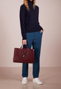 Bordeauxrode leren handtas met dubbele handvatten en een metalen accent, gecombineerd met een marineblauwe coltrui en teal broek, geplaatst tegen een neutrale achtergrond.