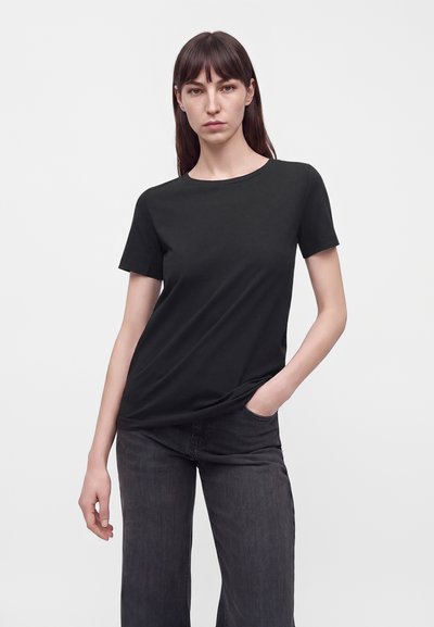 Femme aux cheveux bruns raides portant un t-shirt noir uni à manches courtes et un jean foncé, debout avec une main dans la poche sur fond blanc.
