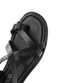Sandalo in pelle nera con design a strap incrociati, texture liscia, suola piatta e dettagli cuciti lungo il bordo.