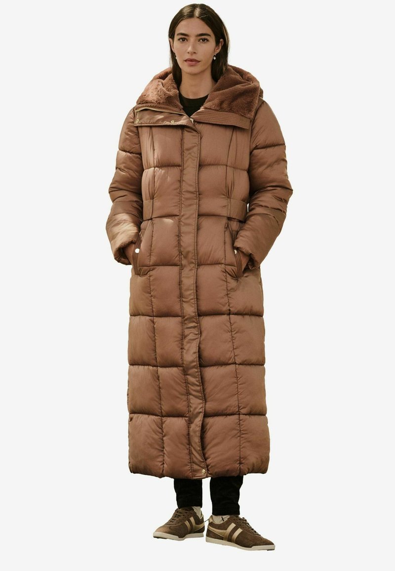 Brun, lang, quiltet pufferjakke med hætte foret med pels, sidelommer og lige snit. Bæret med mørke bukser og brune sneakers.