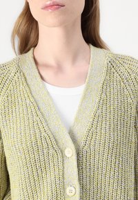 Groen en gele gebreide cardigan met een V-hals ontwerp, voorzien van drie houten knopen en een textuurstof. Daaronder is een wit shirt.
