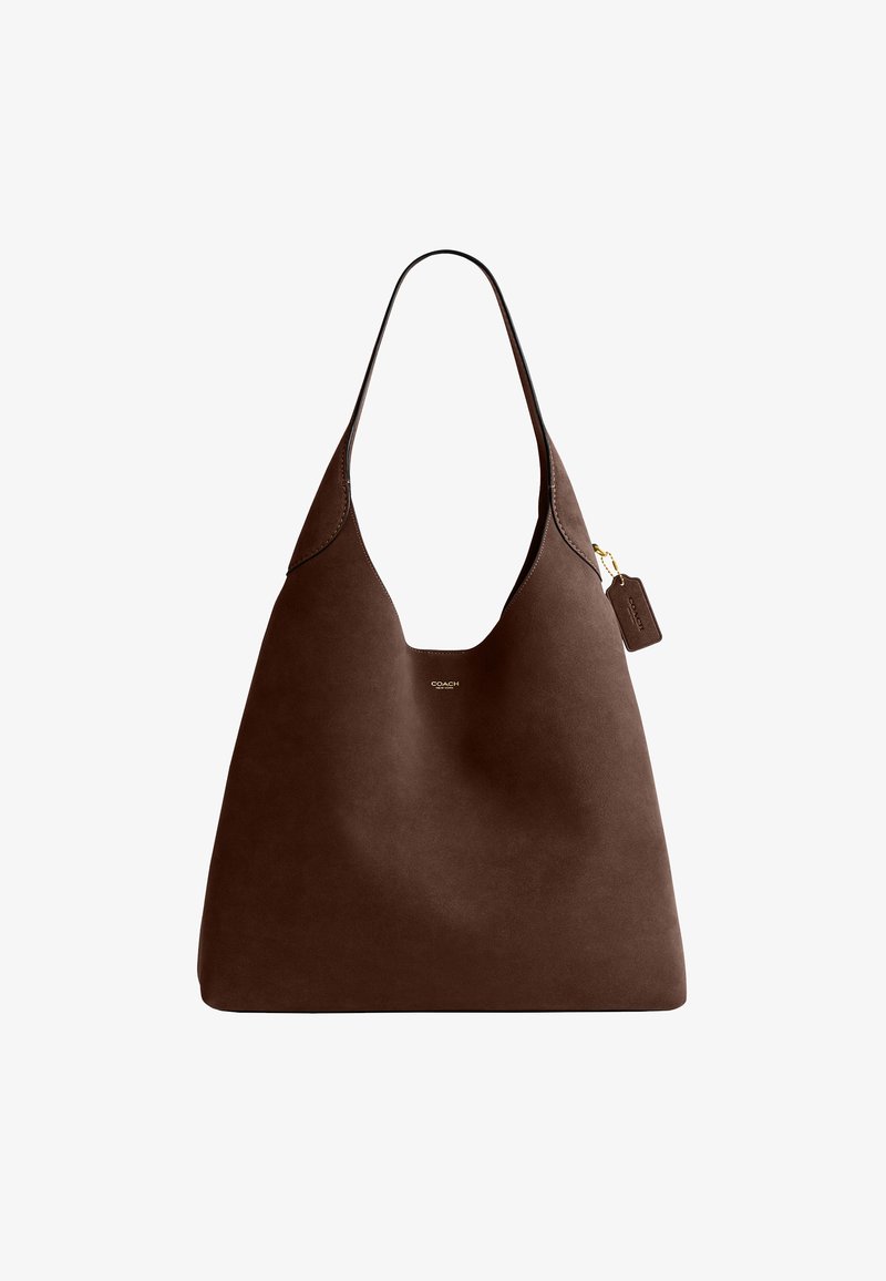 Sac cabas en cuir marron avec une bandoulière unique, petit logo Coach doré et étiquette en cuir attachée sur fond blanc.