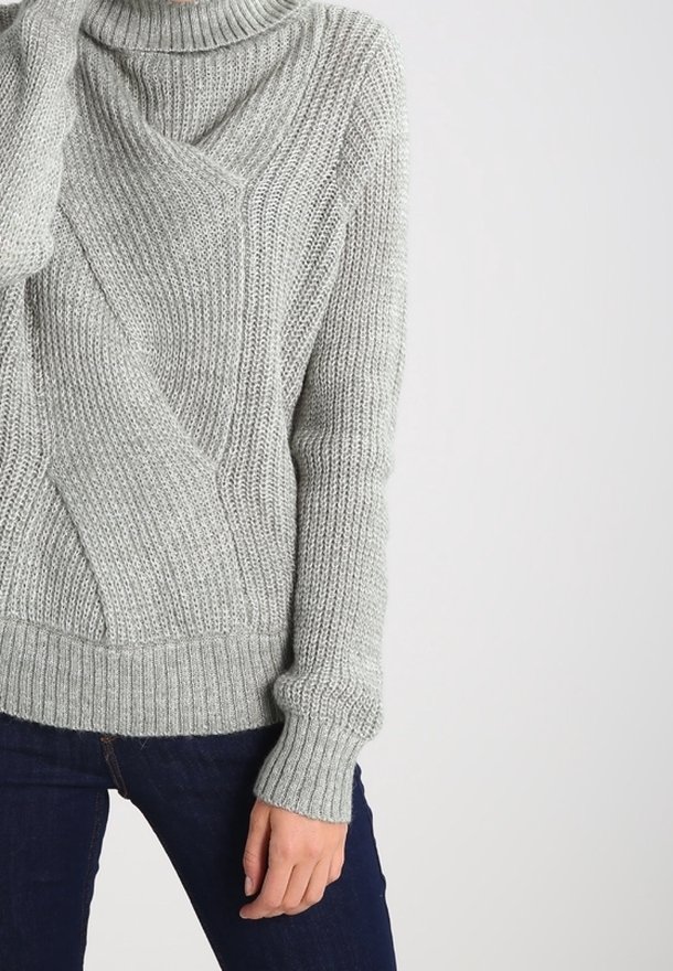 Personne portant un pull en maille épaisse gris clair avec un motif côtelé diagonal et un jean bleu foncé, debout devant un fond clair uni.