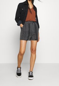 Černá džínová bunda přes hnědý top, spárovaná s vzorovanými černobílými šortkami. Bílé tenisky dokončují outfit.
