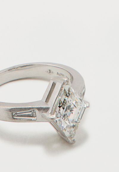 Platina gyűrű, nagy, tiszta, téglalap alakú középső kővel és két kisebb oldalsó kővel. Simított, polírozott felület szögletes dizájnnal.