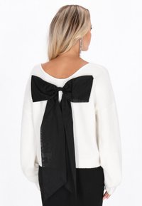 Pulover alb cu detaliu oversized sub formă de fundiță neagră în spate, cu decolteu adânc și mâneci lungi. Textură moale și croi lejer.