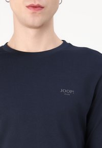 T-shirt in cotone blu navy con scollatura rotonda, caratterizzata da un piccolo logo grigio "JOOP! JEANS" nell'area sinistra del petto. Texture liscia.
