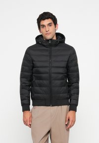 Man wearing a black quilted hooded jacket with zipped pockets and beige pants, standing against a plain white background.