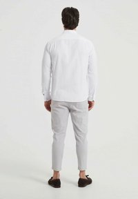 Chemise blanche à manches longues avec col classique, associée à un pantalon slim gris clair et des chaussures brunes, vue de derrière.