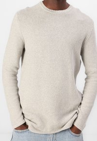 Hellgrauer Strickpullover mit langen Ärmeln, rundem Ausschnitt, strukturiertem Muster und gerippten Bündchen; kombiniert mit hellblauen Jeanshosen.