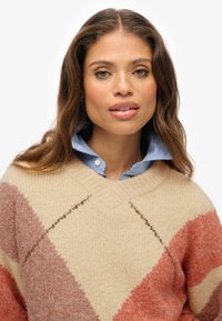 Suéter de mezcla de lana gruesa con una base beige, que presenta patrones de diamantes en color oxidado y durazno. Cuello en V con textura acanalada, superpuesto a una camisa azul.