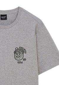 Grijze katoenen t-shirt met een zwarte afbeelding van een leeuw in een tam o' shanter, met de tekst "HIGHLANDER" en "1874" onder de afbeelding.