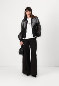 Black mesh bomber jacket with puffed sleeves, white graphic t-shirt, black wide-leg trousers, and a small black handbag.