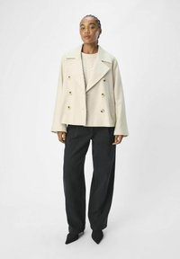 Manteau beige à double boutonnage avec de grands boutons dorés, associé à un léger pull en maille torsadée et un pantalon large et sombre. Texture lisse.