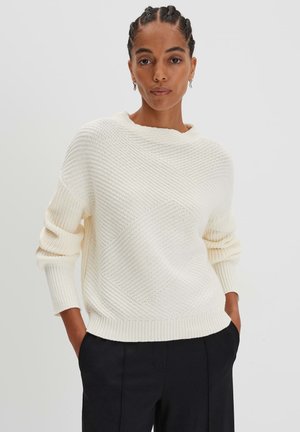 Cream knit sweater with a textured diamond pattern, round neckline, and ribbed cuffs, paired with black trousers.