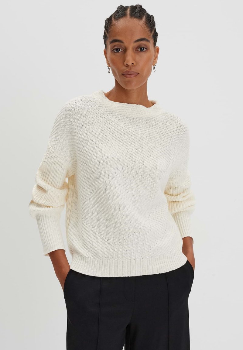 Maglione di maglia color crema con motivo a diamante testurizzato, scollo rotondo e polsini a coste, abbinato a pantaloni neri.