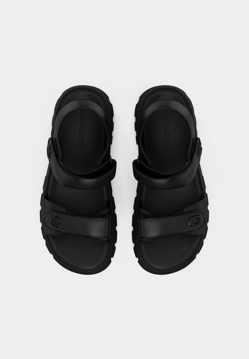 Sandalias de cuero negro con correas ajustables, suelas texturizadas y un logo en relieve. Diseño plano con una estética minimalista.