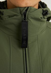 Olivově zelená bunda s vysokým límcem, zipovým zapínáním, černými detaily a visící textilní etiketou s embosovaným textem na přední straně.