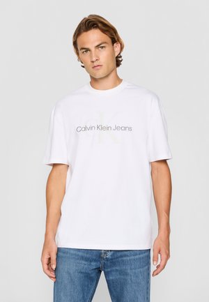 Camiseta de algodón blanca con el logo de "Calvin Klein Jeans" en gris, de mangas cortas y un corte holgado. Combinada con jeans de mezclilla azul.