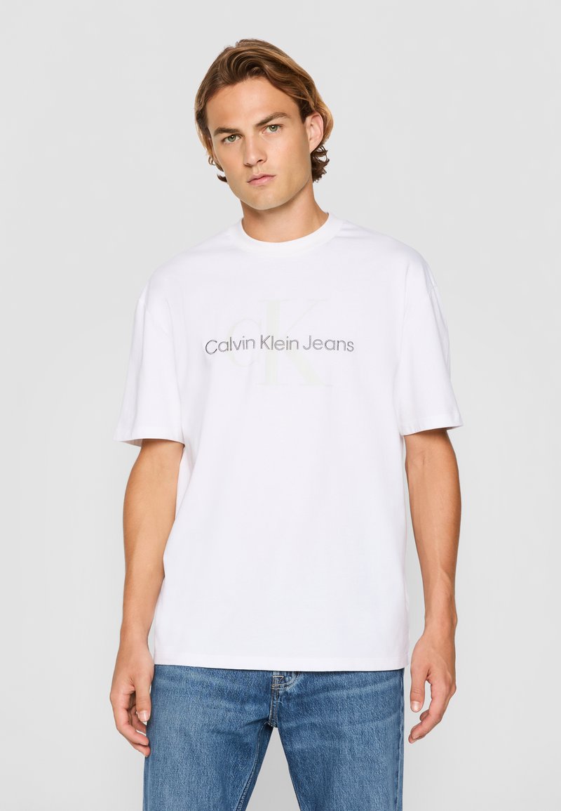 Camiseta de algodón blanca con el logo de "Calvin Klein Jeans" en gris, de mangas cortas y un corte holgado. Combinada con jeans de mezclilla azul.