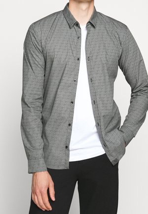 Homme portant une chemise grise à motifs boutonnée sur un t-shirt blanc et un pantalon noir, la main droite dans la poche et la main gauche détendue.