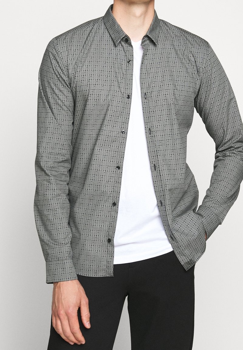 Homme portant une chemise grise à motifs boutonnée sur un t-shirt blanc et un pantalon noir, la main droite dans la poche et la main gauche détendue.