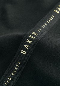 Zwarte stof met een gladde textuur en een geweven label met "BAKER by TED BAKER" in gouden letters langs de rand.