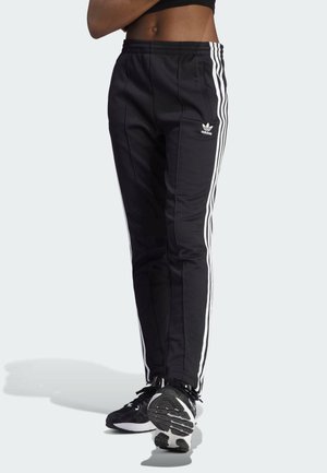 Pantalones de chándal negros de Adidas con rayas blancas a los lados, usados con zapatillas de correr negras y blancas, mostrando la parte inferior del torso y las piernas.