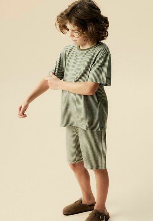 Young child in green-striped short-sleeve shirt and shorts adjusts sleeve, standing on beige background wearing brown slip-on shoes.