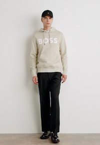 Bežinė hoodie su baltu "BOSS" logotipu, derinama su juodomis kelnėmis ir juodais batais. Modelis dėvi juodą kepurę ir stovi prieš paprastą foną.