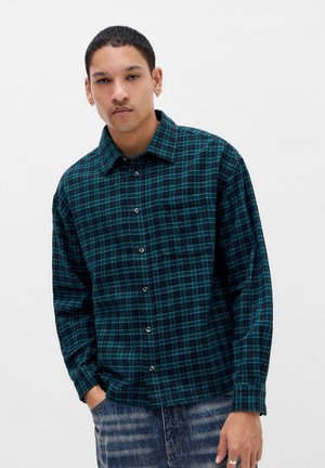 Jeune homme aux cheveux courts porte une chemise à carreaux verts et bleus à boutons et un jean bleu, debout avec une main dans la poche devant un fond blanc.