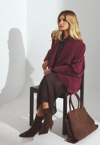 Blazer oversize bordeaux, blouse noire transparente, bottines marron foncé à la cheville et un sac en suède marron, assis sur une chaise noire devant un fond clair.