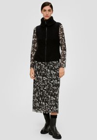 Schwarze Strickweste mit hohem Kragen über einem transparenten, gemusterten Kleid mit grauen und schwarzen Blumenmustern. Knielang, kombiniert mit schwarzen Stiefeln.