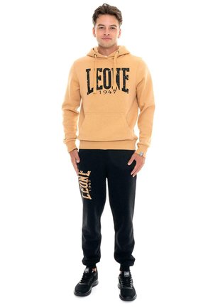 Un hombre posa con una sudadera con capucha Leone 1947 de color beige, pantalones de chándal negros con el logotipo y zapatillas negras sobre un fondo blanco.