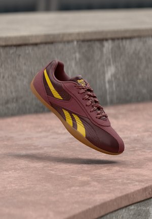 Bordowo-żółty but sportowy Reebok zawieszony nad różowymi kamiennymi schodami na tle szarej kamiennej ściany.