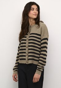 Kootud kardigan pruunis toonis julgete mustade triipudega, ümar kaelus, nööpidega ees ja soonikuäärtega varrukad. Pehme tekstuur ja lühike lõige.