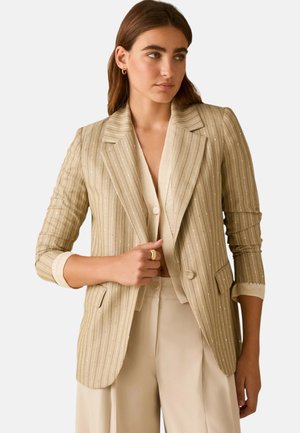 Femme portant un blazer beige à fines rayures sur un top crème boutonné et un pantalon large assorti, debout avec la main près du revers du blazer.