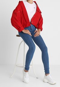 Giacca rossa cropped con maniche lunghe, abbinata a una maglietta bianca e jeans skinny blu. Sneakers bianche completano l'outfit.