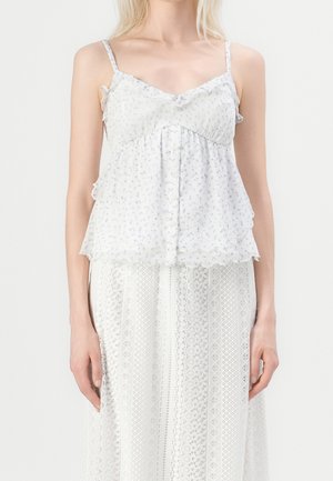 Vrouw draagt een wit mouwloos topje met kleine bloemenprints en een witte kanten rok, staand tegen een effen lichte achtergrond.