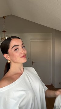 White off-shoulder top with a smooth texture. Model has prominent freckles, gold hoop earrings, and long brown hair styled in a low ponytail.