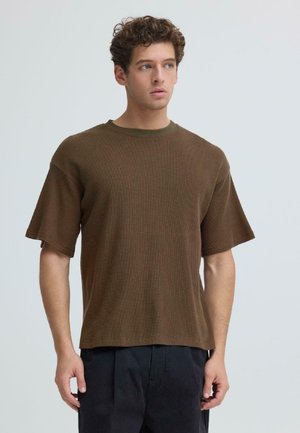 Jonge man met krullend haar, gekleed in een bruin gestructureerd T-shirt met korte mouwen en zwarte broek, kijkt iets opzij tegen een effen achtergrond.