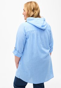 Helles blaues Hoodie-Shirt mit aufgerollten Ärmeln, einem gerafften Rücken und einer lockeren Passform. Verfügt über einen strukturierten, atmungsaktiven Stoff.